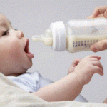 Немовля лежить і відкриває рот, поки дорослий підносить до нього пляшечку з рідиною для годування.