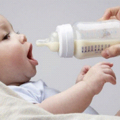 Немовля лежить і відкриває рот, поки дорослий підносить до нього пляшечку з рідиною для годування.