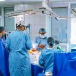 На зображенні показано операційну залу в лікарні. У центрі — пацієнт, який лежить на операційному столі, накритий стерильними простирадлами. Навколо нього працює команда з кількох медичних працівників — хірурги та асистенти. Вони одягнені у блакитні стерильні халати, шапочки та маски, що закривають обличчя. Над столом розташовані великі операційні лампи, які яскраво освітлюють робочу зону. Медики зосереджені на проведенні хірургічного втручання: їхні руки спрямовані до операційного поля, поруч видно медичні інструменти та обладнання. Загальна атмосфера — стерильна, професійна та напружена, характерна для проведення складної операції.