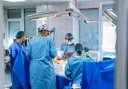 На зображенні показано операційну залу в лікарні. У центрі — пацієнт, який лежить на операційному столі, накритий стерильними простирадлами. Навколо нього працює команда з кількох медичних працівників — хірурги та асистенти. Вони одягнені у блакитні стерильні халати, шапочки та маски, що закривають обличчя. Над столом розташовані великі операційні лампи, які яскраво освітлюють робочу зону. Медики зосереджені на проведенні хірургічного втручання: їхні руки спрямовані до операційного поля, поруч видно медичні інструменти та обладнання. Загальна атмосфера — стерильна, професійна та напружена, характерна для проведення складної операції.