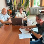 Три людини за столом: жінка з ноутбуком та двоє чоловіків. На задньому плані український прапор і військові артефакти