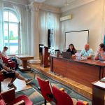 Актова зала в районній військові адміністрації, де троє людей сидять за столом, а двоє – в залі