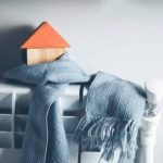 Маленький дерев'яний будиночок з помаранчевим дахом, обгорнутий синім шарфом, стоїть на білому радіаторі