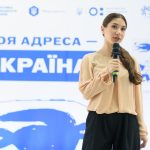 Молода жінка говорить у мікрофон на тлі банера з текстом "ТВОЯ АДРЕСА УКРАЇНА" та синіми зображеннями