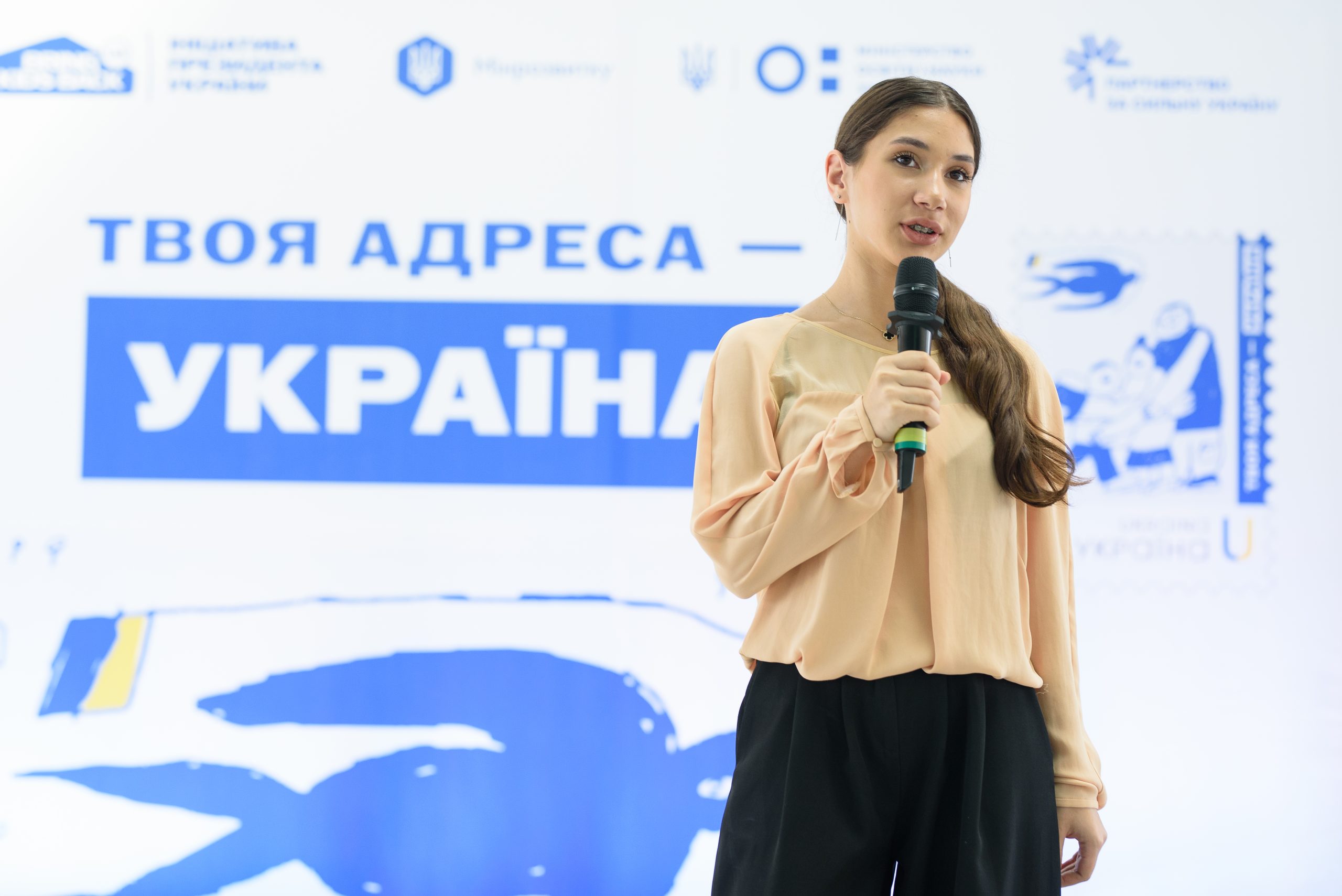 Молода жінка говорить у мікрофон на тлі банера з текстом "ТВОЯ АДРЕСА УКРАЇНА" та синіми зображеннями