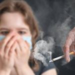 Рука тримає димлячу сигарету, а на тлі розмита людина в масці закриває ніс і рот