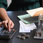 Людина використовує калькулятор поруч з документами, монетами, банкою для грошей та моделлю будинку, що натякає на фінансове планування