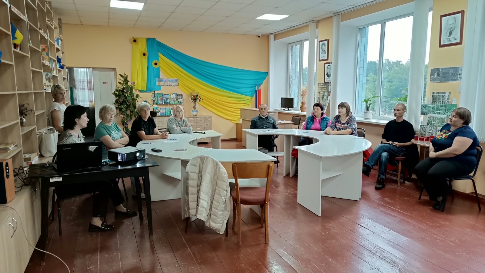 Група людей сидить за півколом білих столів під час зустрічі в бібліотеці з жовто-блакитним прапором на стіні